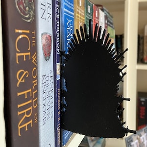 May include: A black metal bookend shaped like the Iron Throne from the Game of Thrones series. The bookend is placed between several books with titles including "The Ice Dragon" and "A Knight of the Seven Kingdoms". The books are on a white shelf.