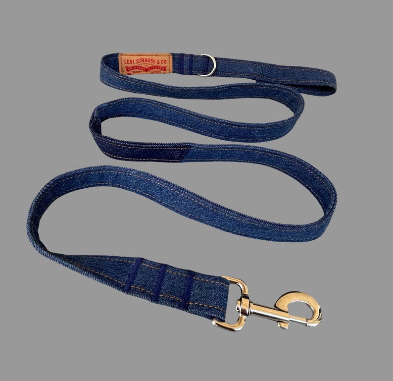 May include: A blue denim dog leash and matching collar. The leash has a silver-colored clasp and a loop handle. The collar features a Levi Strauss & Co. label. The leash and collar are made of blue denim fabric with visible stitching.