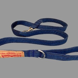 May include: A blue denim dog leash and matching handle. The leash features a silver metal clasp and a silver metal ring. A brown leather patch with the text "LEVI STRAUSS & CO." is sewn onto the leash.