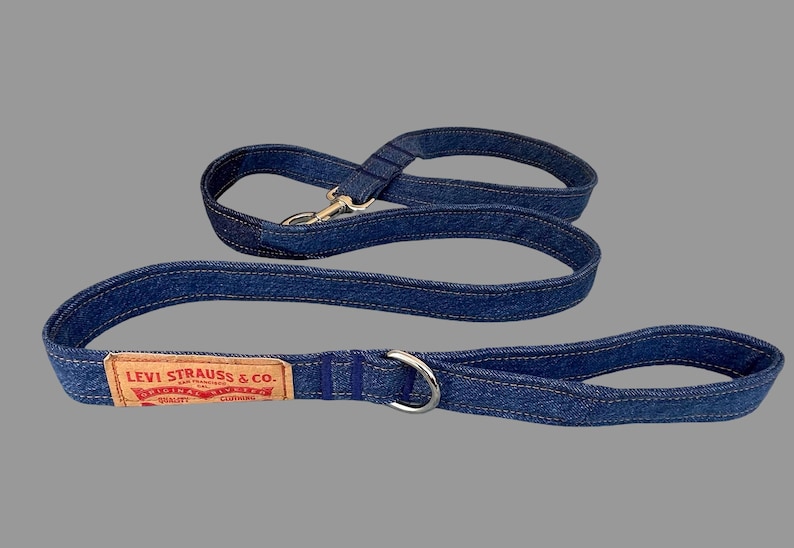 May include: A blue denim dog leash and matching handle. The leash features a silver metal clasp and a silver metal ring. A brown leather patch with the text "LEVI STRAUSS & CO." is sewn onto the leash.
