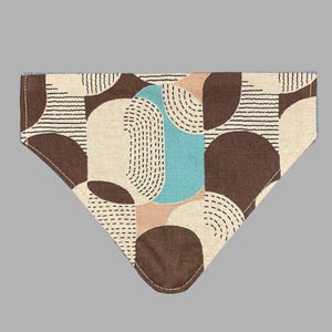 K&ouml;nnte beinhalten: Dreieckiges Haustier-Bandana mit geometrischem Muster. Das Design zeigt braune, beige und hellblaue Formen mit schwarzen gestrichelten Linien und horizontalen Streifen. Die Kanten sind mit wei&szlig;em Faden gen&auml;ht.
