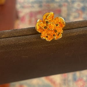 May include: A gold-toned metal lapel pin shaped like a five-petaled flower. The surface of the flower features a textured, raised design with small, dark, circular accents embedded within the center and petals, creating a detailed floral appearance.