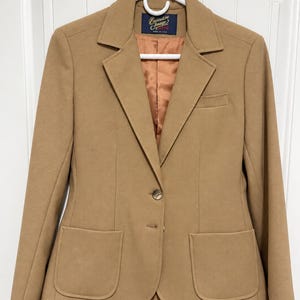 May include: A tan blazer featuring a notched lapel, two front patch pockets, and a single chest pocket. The garment has a two-button closure and a smooth, copper-coloured interior lining. A dark blue label inside reads Executive Image Made in USA.