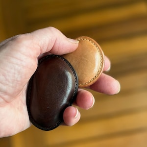 May include: Two leather coasters in hand. One is a dark brown, the other is tan. Both are oval-shaped with stitched edges. The coasters are made of a smooth, polished leather.