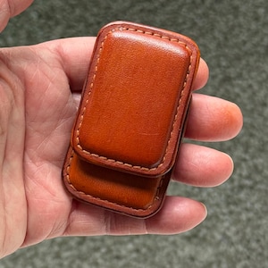 May include: A rectangular, brown leather money clip with a magnetic closure. The clip is made of smooth, stitched leather and is held in a hand. The item is designed to hold cash and cards.