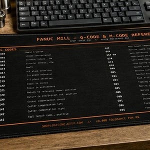 Könnte beinhalten: Eine schwarze, rechteckige Schreibtischunterlage mit orangefarbenem Text und Linien. Die Unterlage zeigt eine Fanuc Mill G-Code und M-Code Referenz mit Listen von Codes und deren Funktionen sowie das ShopLogic CNC Logo oben links.