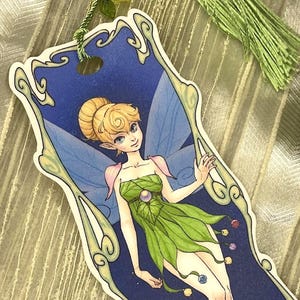 May include: A bookmark featuring a whimsical illustration of a fairy with blonde hair, green wings, and a green dress. The fairy is surrounded by a blue background with a decorative frame.
