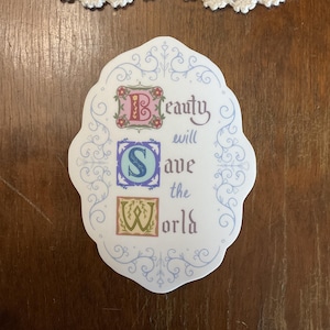 May include: A white sticker with a blue, ornate frame. The words "Beauty will save the world" are written in a decorative font with the letters B, S, and W in colorful squares.