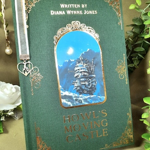 May include: A green hardcover book with gold accents and a white ribbon bookmark. The book cover features an illustration of a flying castle against a blue sky with a moon and stars. The title "Howl's Moving Castle" is written in gold lettering on the front cover. The book is by Diana Wynne Jones.