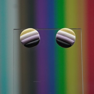 Könnte beinhalten: Ein Paar runde Ohrringe mit einem gestreiften Design in Gelb, Weiß, Lila und Schwarz. Die Ohrringe werden auf einer durchsichtigen Oberfläche vor einem verschwommenen Regenbogenhintergrund gezeigt.