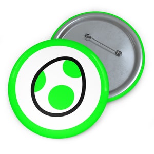 May include: A green and white button with a black outline of a green egg with three white dots. The button has a metal back.
