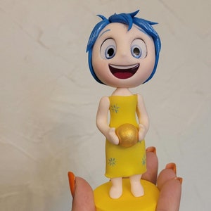 Könnte beinhalten: Eine gelbe Figur von Freude aus dem Film Inside Out. Freude hält einen goldenen Ball in ihren Händen. Sie trägt ein gelbes Kleid mit weißen Sternen. Die Figur steht auf einem gelben Sockel.