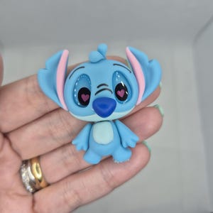 Puede incluir: Una figura azul del personaje Stitch de la pel&iacute;cula de Disney Lilo & Stitch. La figura tiene orejas grandes con el interior rosa, ojos grandes y una nariz peque&ntilde;a. La figura est&aacute; posando con los brazos a los lados.
