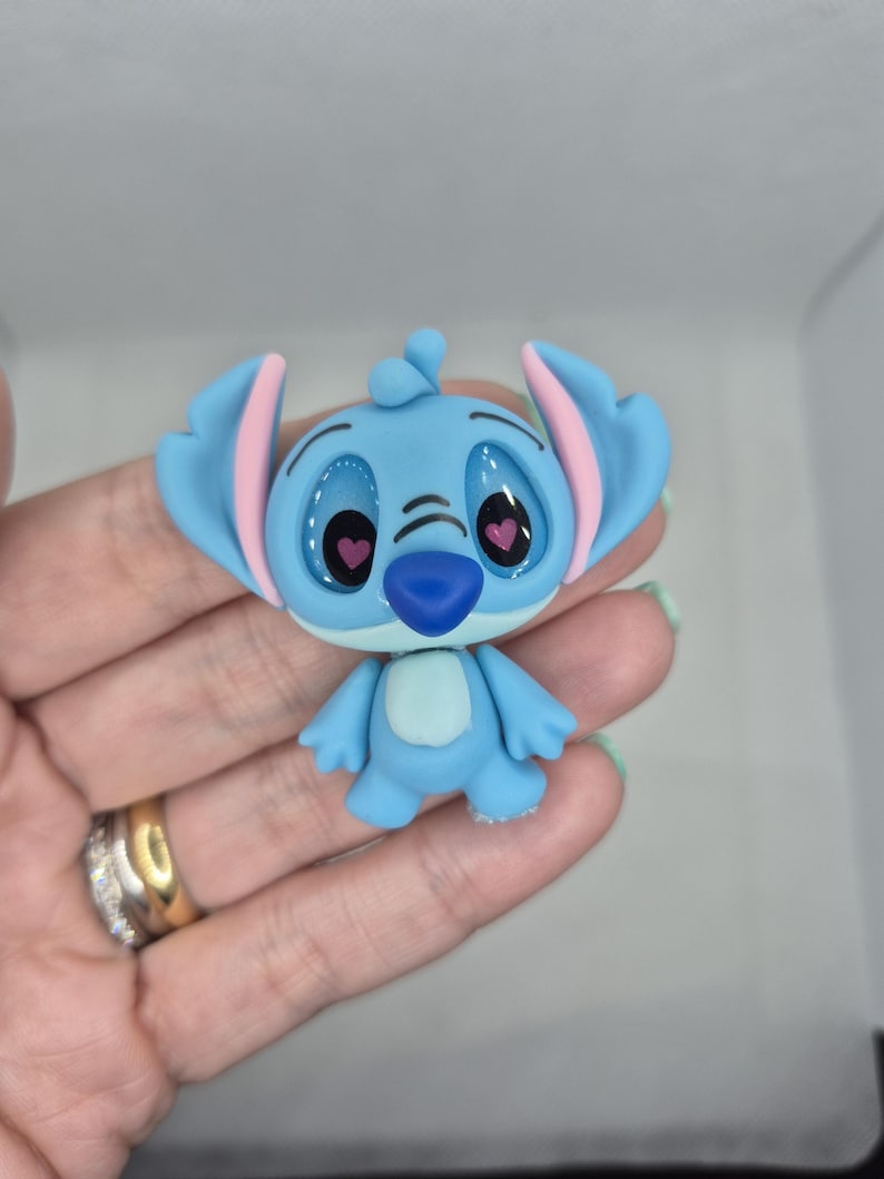 Puede incluir: Una figura azul del personaje Stitch de la pel&iacute;cula de Disney Lilo & Stitch. La figura tiene orejas grandes con el interior rosa, ojos grandes y una nariz peque&ntilde;a. La figura est&aacute; posando con los brazos a los lados.