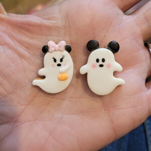 Puede incluir: Dos galletas blancas en forma de fantasma con ojos negros y mejillas rosadas. Un fantasma tiene un lazo rosa en la cabeza y el otro tiene orejas negras de Mickey Mouse.