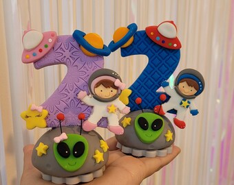 以下が含まれることがあります： 宇宙をテーマにしたパターンで飾られた、紫と青の数字のケーキトッパー。宇宙飛行士と宇宙人のフィギュアが付いています。