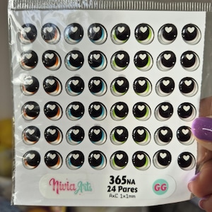 Op de afbeelding: Een vel met 24 paar zwarte, witte en kleurrijke plastic poppenogen met een hartvorm in het midden. Het vel is gelabeld met "365NA 24 Pares AxL 1x1mm" en "GG".