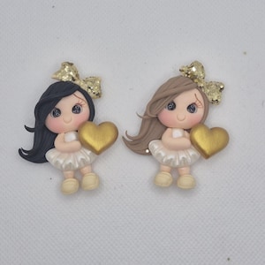 May include: Two doll figures, each holding a gold heart. One doll has black hair, the other has brown hair. Both dolls wear white dresses, beige shoes, and gold glitter bows.