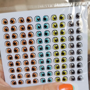 Op de afbeelding: Een vel met kleurrijke knuffelstickers. Het vel heeft 7 rijen met elk 6 ogen. De ogen zijn in 4 kleuren: oranje, blauw, geel en grijs.