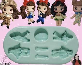 Puede incluir: Un molde de silicona azul claro con varias formas de muñecas, incluyendo una muñeca sosteniendo un perro, y varias figuras de muñecas con lazos. El molde está sobre un fondo rosa con la palabra "Artesanato" en la esquina inferior izquierda.