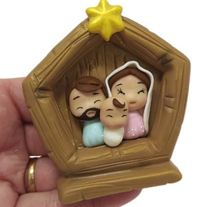 Puede incluir: Un pequeño pesebre de madera de color dorado con una estrella en la parte superior. Dentro de la escena hay tres figuras: un hombre con barba, una mujer y un bebé. Las figuras están hechas de arcilla y tienen un estilo de dibujos animados.