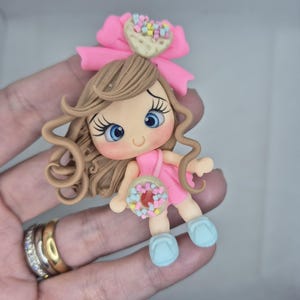 Könnte beinhalten: Handgefertigte Polymer Clay Figur einer Mädchenfigur in rosa Kleidung, mit blauen Augen und braunen Haaren. Die Figur hat eine rosa Schleife mit herzförmiger Dekoration und hält einen Mini-Donut. Die Schuhe sind hellblau.