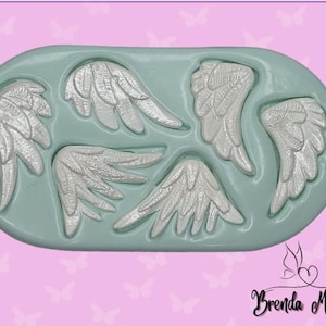 May include: A light teal silicone mold with six angel wing designs in various sizes. The wings are detailed with feather textures and are a light silver color. The mold is oval-shaped and sits on a pink background with butterfly graphics.