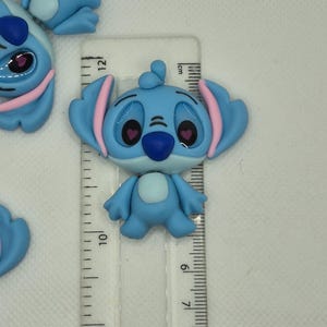 Puede incluir: Figura de Stitch azul claro con detalles rosas, de aproximadamente 5 cm de altura. La figura tiene orejas grandes, una nariz azul oscuro y ojos en forma de coraz&oacute;n. Una regla transparente con marcas de cent&iacute;metros est&aacute; al lado.