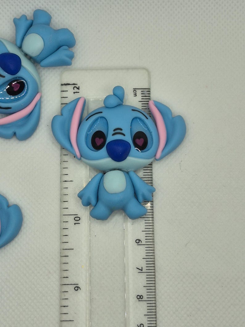 Puede incluir: Figura de Stitch azul claro con detalles rosas, de aproximadamente 5 cm de altura. La figura tiene orejas grandes, una nariz azul oscuro y ojos en forma de coraz&oacute;n. Una regla transparente con marcas de cent&iacute;metros est&aacute; al lado.
