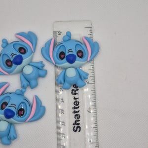 Puede incluir: Varios dijes del personaje Stitch de color azul claro con detalles en rosa, colocados cerca de una regla. Los dijes tienen orejas grandes, narices azul oscuro y ojos rosas. La regla est&aacute; marcada en cent&iacute;metros.