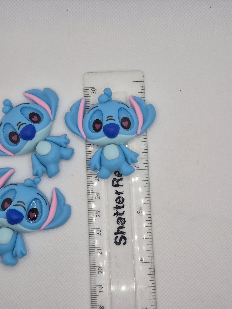Puede incluir: Varios dijes del personaje Stitch de color azul claro con detalles en rosa, colocados cerca de una regla. Los dijes tienen orejas grandes, narices azul oscuro y ojos rosas. La regla est&aacute; marcada en cent&iacute;metros.