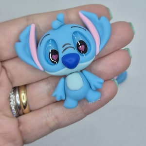 Puede incluir: Figura azul claro de un personaje de dibujos animados con orejas grandes, interior de las orejas rosa y nariz azul. El personaje tiene ojos grandes con pupilas rosas en forma de coraz&oacute;n. La figura se sostiene en una mano.