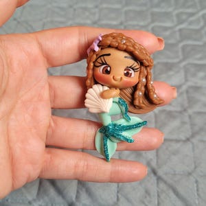 May include: A brown-skinned mermaid doll with brown hair, a seashell, and a teal tail with glitter. The doll is wearing a light green top.
