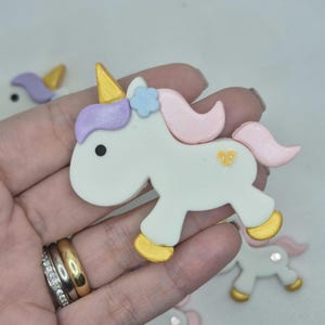 May include: A white unicorn figurine with a gold horn, hooves, and a heart-shaped detail. The unicorn has a pink mane and tail, a light blue flower, and a purple ear. Additional unicorn figurines are visible in the background.