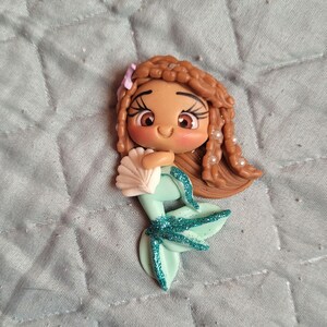 May include: A brown-skinned mermaid doll with brown hair, a green tail with glitter, and a white seashell. The doll is smiling and has large brown eyes.