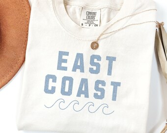 May include: A cream-coloured t-shirt with the text "EAST COAST" in blue lettering and a row of blue waves underneath. The shirt is folded and has a gold necklace with a pendant on top.