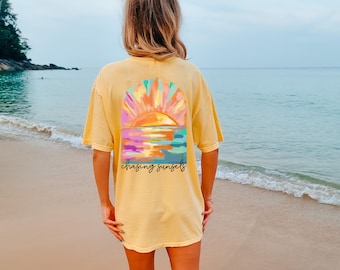 May include: Yellow t-shirt with a colorful sunset graphic on the back. The design features a sun over the ocean with the words "chasing sunsets" in a script font. The shirt is worn on a beach.