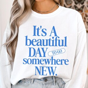 K&ouml;nnte beinhalten: Wei&szlig;es Langarm-Sweatshirt mit blauem Text: "It's A beautiful DAY to go somewhere NEW." Das l&auml;ssige Sweatshirt ist f&uuml;r den Alltag geeignet. Der Text ist in einer fetten, serifenlosen Schriftart.