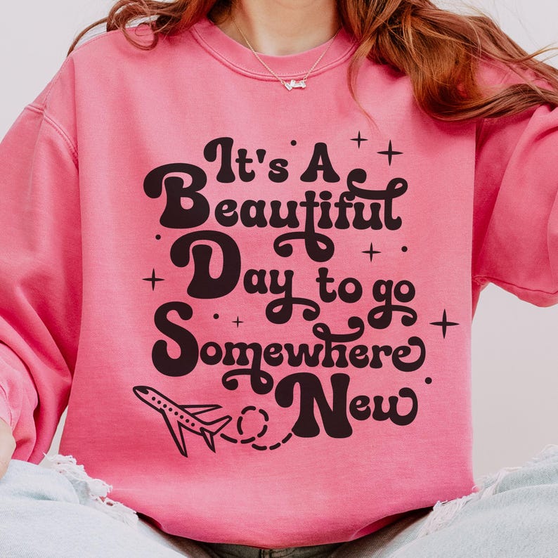 以下が含まれることがあります： 「It's A Beautiful Day to go Somewhere New」の黒い文字がプリントされたピンク色のスウェットシャツ。テキストの下には小さな飛行機のグラフィックがあります。スウェットシャツは無地のピンク色です。
