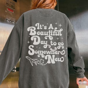 以下が含まれることがあります： 「It's A Beautiful Day to go Somewhere New」の白文字と飛行機のグラフィックがプリントされた濃いグレーのスウェットシャツ。 ヴィンテージ感のあるウォッシュ加工が施されています。