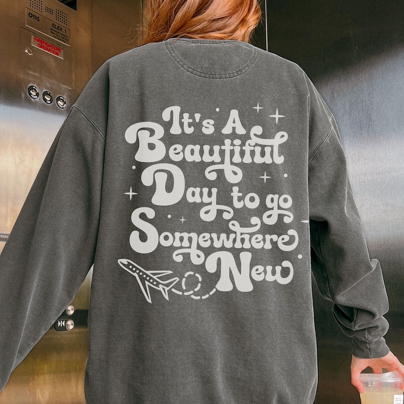 以下が含まれることがあります： 「It's A Beautiful Day to go Somewhere New」の白文字と飛行機のグラフィックがプリントされた濃いグレーのスウェットシャツ。 ヴィンテージ感のあるウォッシュ加工が施されています。
