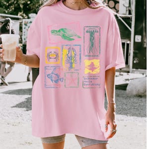 May include: Pink t-shirt featuring a collection of sea creatures in colorful frames. The design includes a turtle, jellyfish, crab, lobster, fish, starfish, and the text "Saltwater Heals Everything."