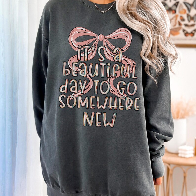 Puede incluir: Sudadera gris oscuro con el texto "It's a beautiful day to go somewhere new" en una paleta de colores pastel. El texto est&aacute; superpuesto a un gr&aacute;fico de lazo rosa. La sudadera es de corte holgado.