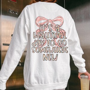 Puede incluir: Sudadera blanca con un lazo rosa y el texto "It's a beautiful day to go somewhere new". La sudadera tiene mangas largas y cuello redondo. El gr&aacute;fico est&aacute; en una paleta de colores pastel.