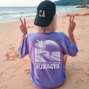 May include: A purple t-shirt with a white graphic of a palm tree on a beach with the text "Chasing Sunsets" printed below the graphic. The person wearing the shirt is facing away from the camera and has a black baseball cap with the letters "CA" on the front.