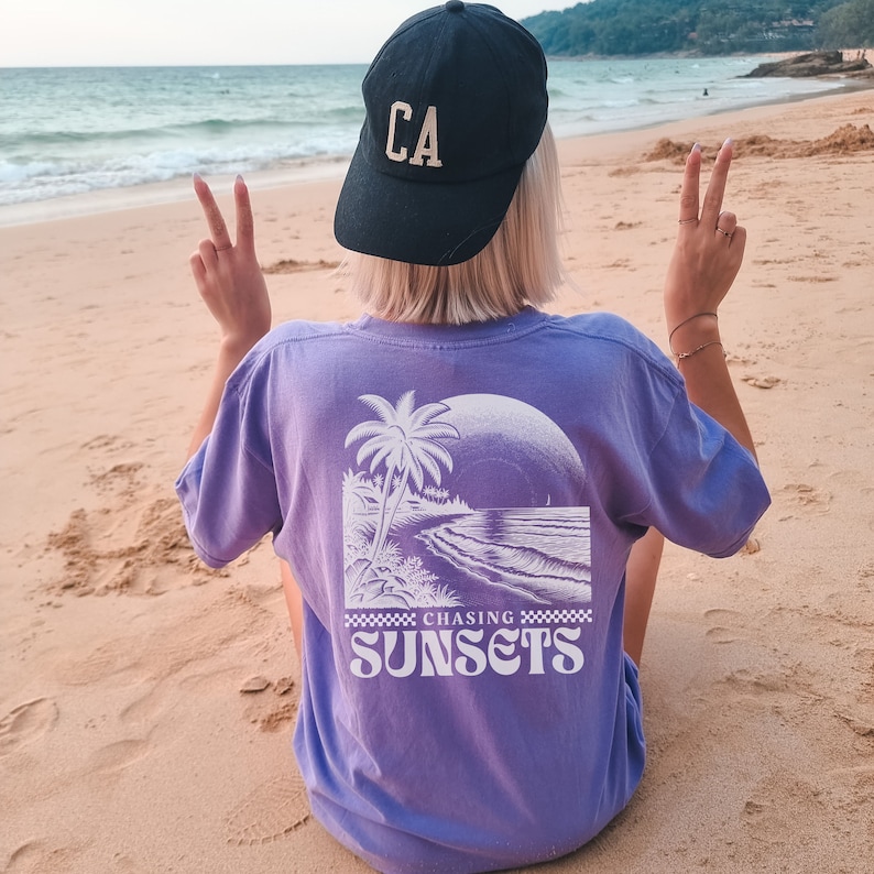 May include: A purple t-shirt with a white graphic of a palm tree on a beach with the text "Chasing Sunsets" printed below the graphic. The person wearing the shirt is facing away from the camera and has a black baseball cap with the letters "CA" on the front.