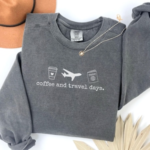 Puede incluir: Una sudadera gris carb&oacute;n con las palabras "coffee and travel days" impresas en blanco. El dise&ntilde;o incluye una taza de caf&eacute;, un avi&oacute;n y un pasaporte. Tambi&eacute;n se ven un collar dorado y un sombrero marr&oacute;n.