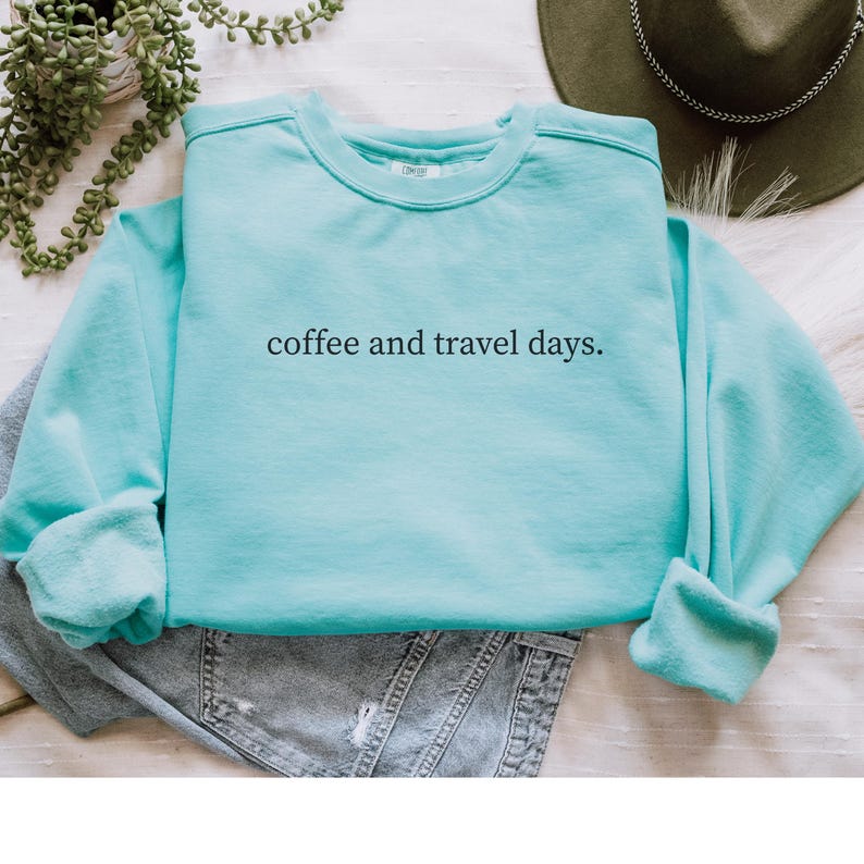 May include: A light blue sweatshirt with the text "coffee and travel days." The sweatshirt is folded on top of a pair of gray jeans. A green hat and a plant are visible in the background.