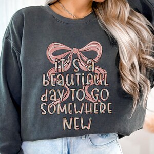 Puede incluir: Sudadera gris oscuro con el texto "IT'S A BEAUTIFUL DAY TO GO SOMEWHERE NEW" en una fuente colorida y caprichosa. Un gran lazo rosa est&aacute; encima del texto. La sudadera es de estilo casual.