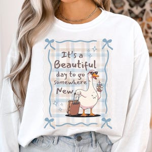 May include: White long-sleeved sweatshirt with a graphic of a goose wearing sunglasses and holding a drink and suitcase. The text reads "It's a Beautiful day to go somewhere New."